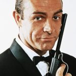 A photograph of Roger Moore as James Bond holding a gun and looking directly at the camera
