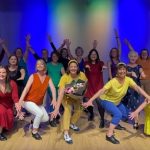 A group of 17 adults in colourful clothes pose for the camera with their arms in the air
