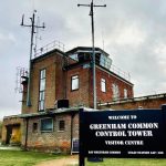 A photo of Greenham Control Tower with a sign reading "Welcome to Greenham Control Tower visitor centre. RAF Greenham Common. USAAF Station AAF-486"