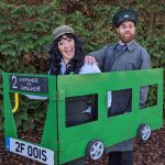 A man and a woman wearing old-fashioned bus conductor outfits inside a green cardboard bus