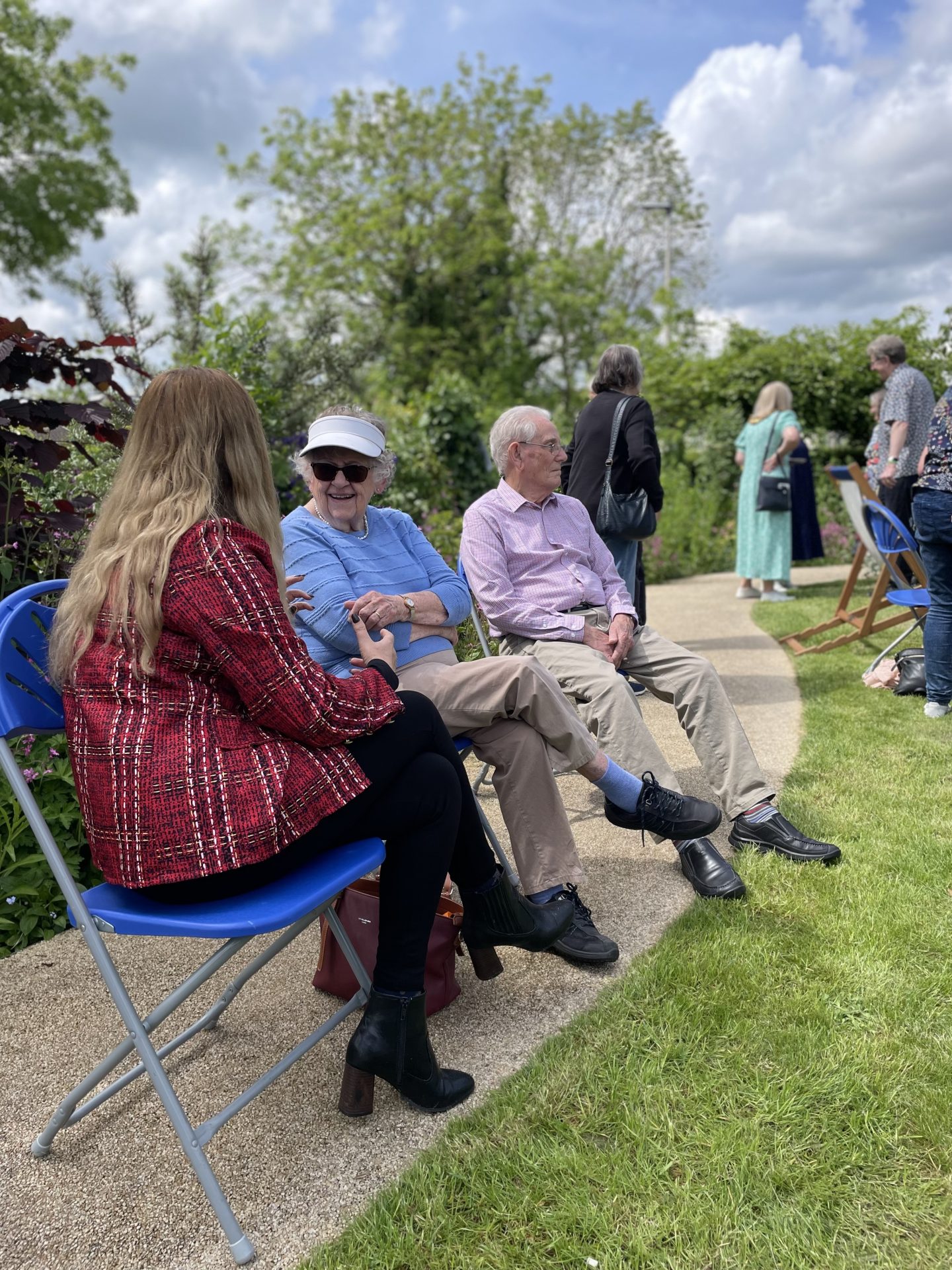 The image shows an outdoor gathering in a garden setting on a sunny day. Three people are seated on blue folding chairs in the foreground. An older woman wearing sunglasses, a white visor, and a blue sweater smiles and chats with a younger woman with long, blonde hair dressed in a red plaid jacket. Next to them, an older man wearing glasses, a light purple shirt, and beige pants sits quietly. Behind them, other people stand and walk along a pathway surrounded by greenery and trees, enjoying the garden and the pleasant weather. The atmosphere appears relaxed and social.