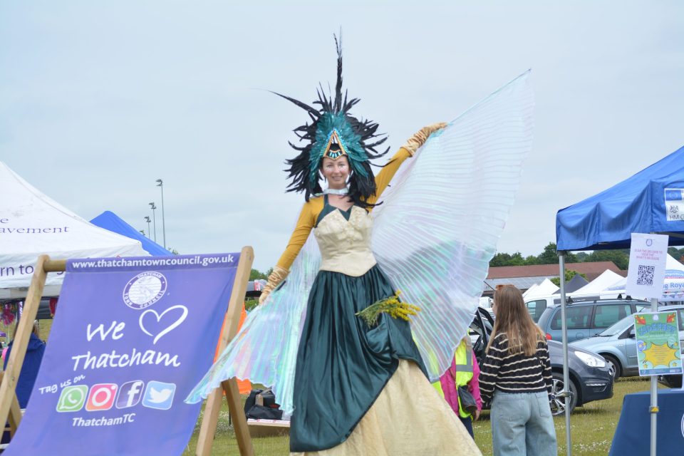 A Stilt walker wearing a green and cream dress and Opaque wings. next to Thatcham Town Council Deck Chair.