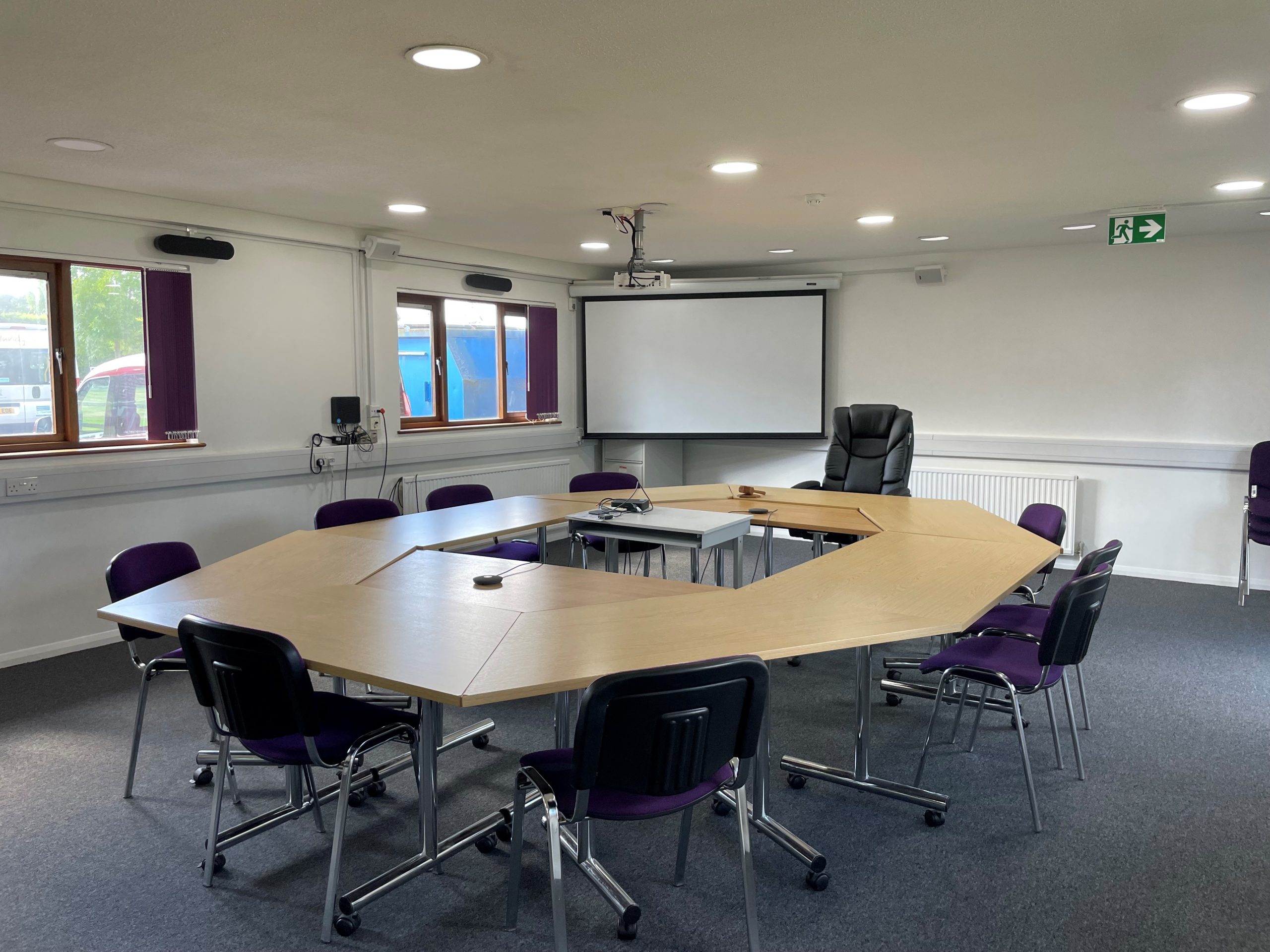 A spacious meeting room with a large, O-shaped table arrangement surrounded by office chairs. The room is well-lit with natural light from several windows and features ceiling-mounted lighting. A projector is installed on the ceiling, with additional office furniture and chairs arranged along the walls. The room has a modern, professional feel, suitable for meetings or events.