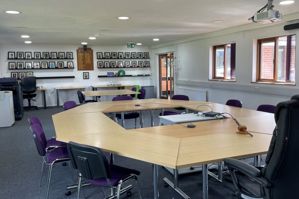 A spacious meeting room with a large, U-shaped table arrangement surrounded by office chairs. The room is well-lit with natural light from several windows and features ceiling-mounted lighting. A projector is installed on the ceiling, with additional office furniture and chairs arranged along the walls. There are framed portraits displayed on one wall, and large windows offer views of green outdoor space. The room has a modern, professional feel, suitable for meetings or events.