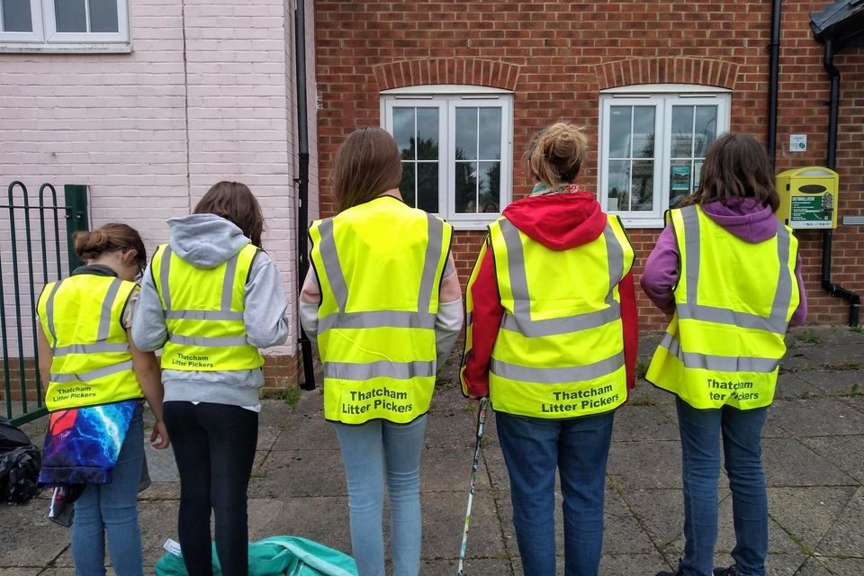 Five people, likely teenagers, are standing with their backs to the camera, wearing bright yellow high-visibility vests with the words 'Thatcham Litter Pickers' printed on the back. They are standing in front of a brick building with two windows. Some of them are holding litter-picking tools, and there is a bag on the ground to their left