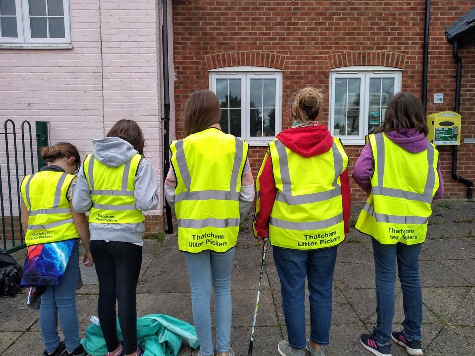 Five people, likely teenagers, are standing with their backs to the camera, wearing bright yellow high-visibility vests with the words 'Thatcham Litter Pickers' printed on the back. They are standing in front of a brick building with two windows. Some of them are holding litter-picking tools, and there is a bag on the ground to their left