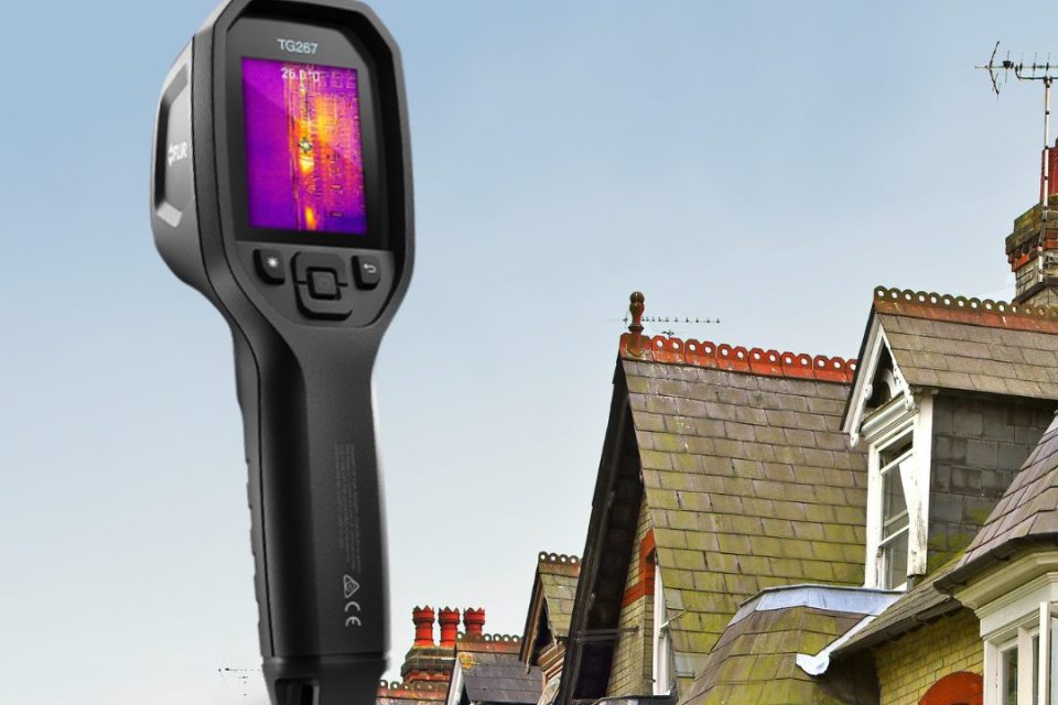 The image shows a handheld thermal imaging camera labeled "TG267" prominently in the foreground. The device’s screen displays a thermal heat map with bright yellow and purple colors indicating temperature variations. In the background, there are a row of traditional British-style houses with steep, slate-tiled roofs and red brick chimneys.