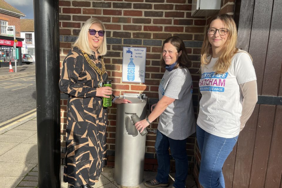 This image shows three women standing near a public water refill station mounted on a brick wall. The woman on the left, wearing a patterned black and beige dress with a ceremonial gold chain around her neck, is holding a green reusable bottle. The woman in the centre, wearing a grey T-shirt, is using the water station to refill a bottle. The woman on the right, dressed in a white T-shirt with the text “Plastic Free Thatcham” and “Surfers Against Sewage” printed on it, stands smiling beside them. The background shows a brick wall with a sign indicating free water refills.