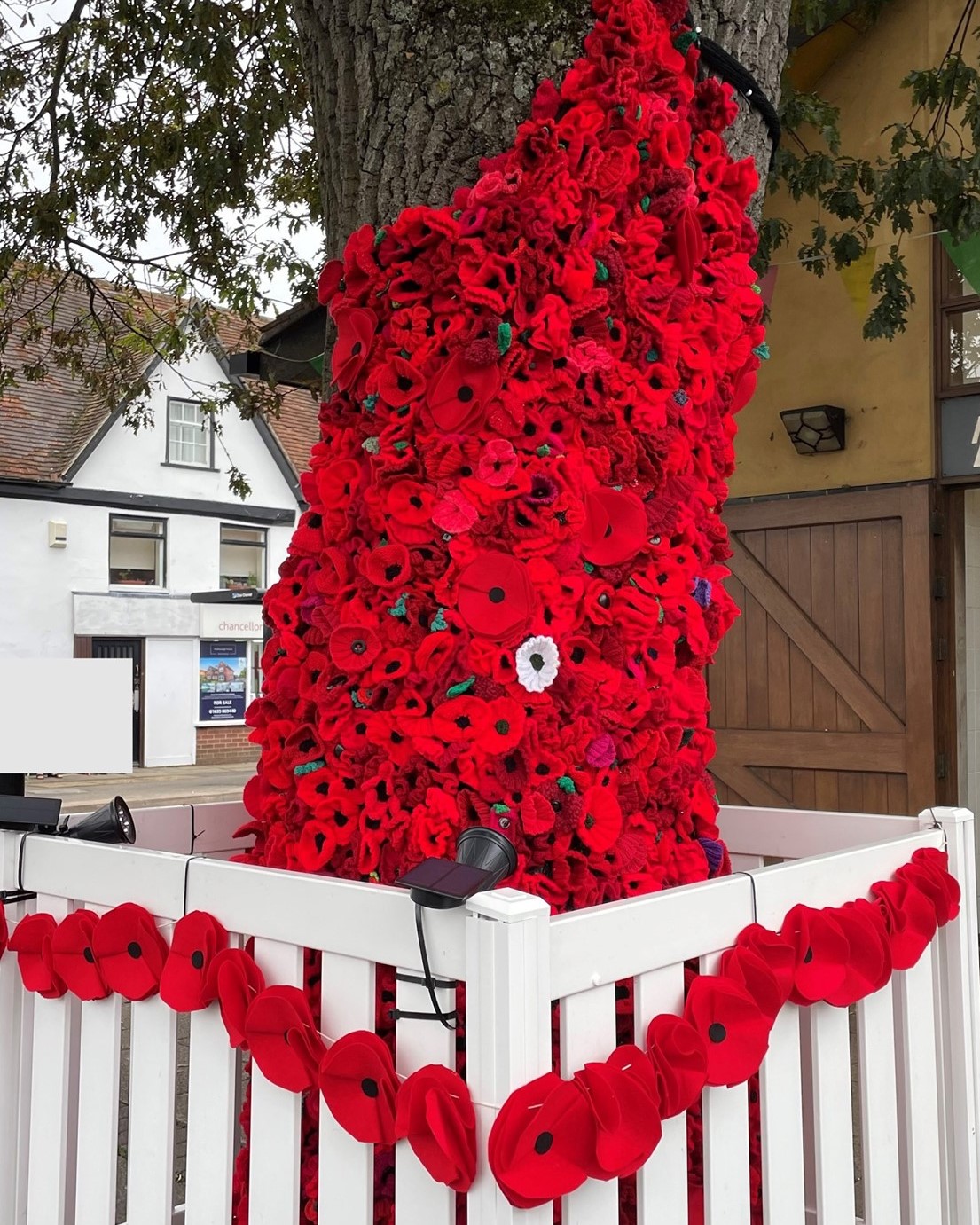 The image shows a tree trunk wrapped in a thick cascade of large, handmade red poppies. The poppies are densely packed, with a single white one standing out among them. A white picket fence surrounds the base of the tree, with a garland of more red poppies draped across the fence.