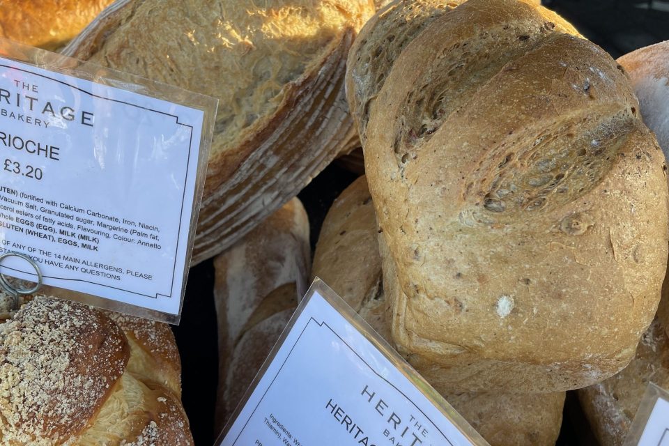 A market stall displaying various types of fresh bread from "The Heritage Bakery." Visible loaves include a crusty "Heritage Sourdough" priced at £4.80, a braided "Brioche" loaf at £3.20 topped with crumbs, and a "Heritage Baguette" priced at £2.20. Each item has a descriptive sign listing ingredients and allergy information. The bread appears golden and freshly baked.