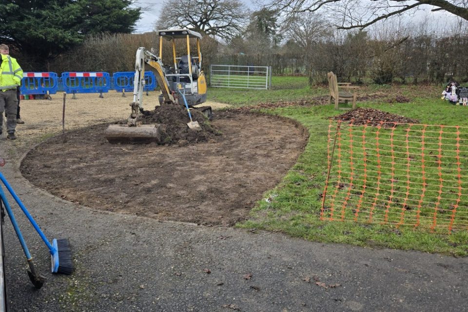 A construction site in a grassy area shows a small yellow excavator digging soil for a new memorial space. The ground has been cleared and leveled, forming a curved shape. A worker in a high-visibility jacket stands nearby. Orange safety netting and blue barriers surround the site, and a wooden bench sits beneath a large tree.