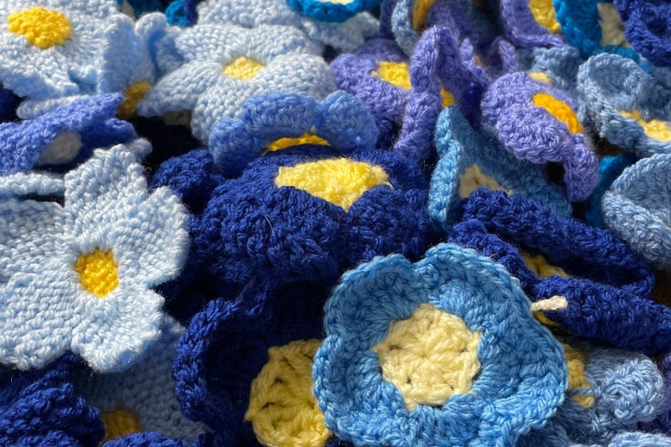 A collection of crocheted flowers in various shades of blue with yellow centres. The flowers are made of yarn and have a soft, textured appearance. The variety of blue hues—from light sky blue to deep navy—adds depth and a sense of richness to the composition. It's a lovely and cosy display of fibre art!