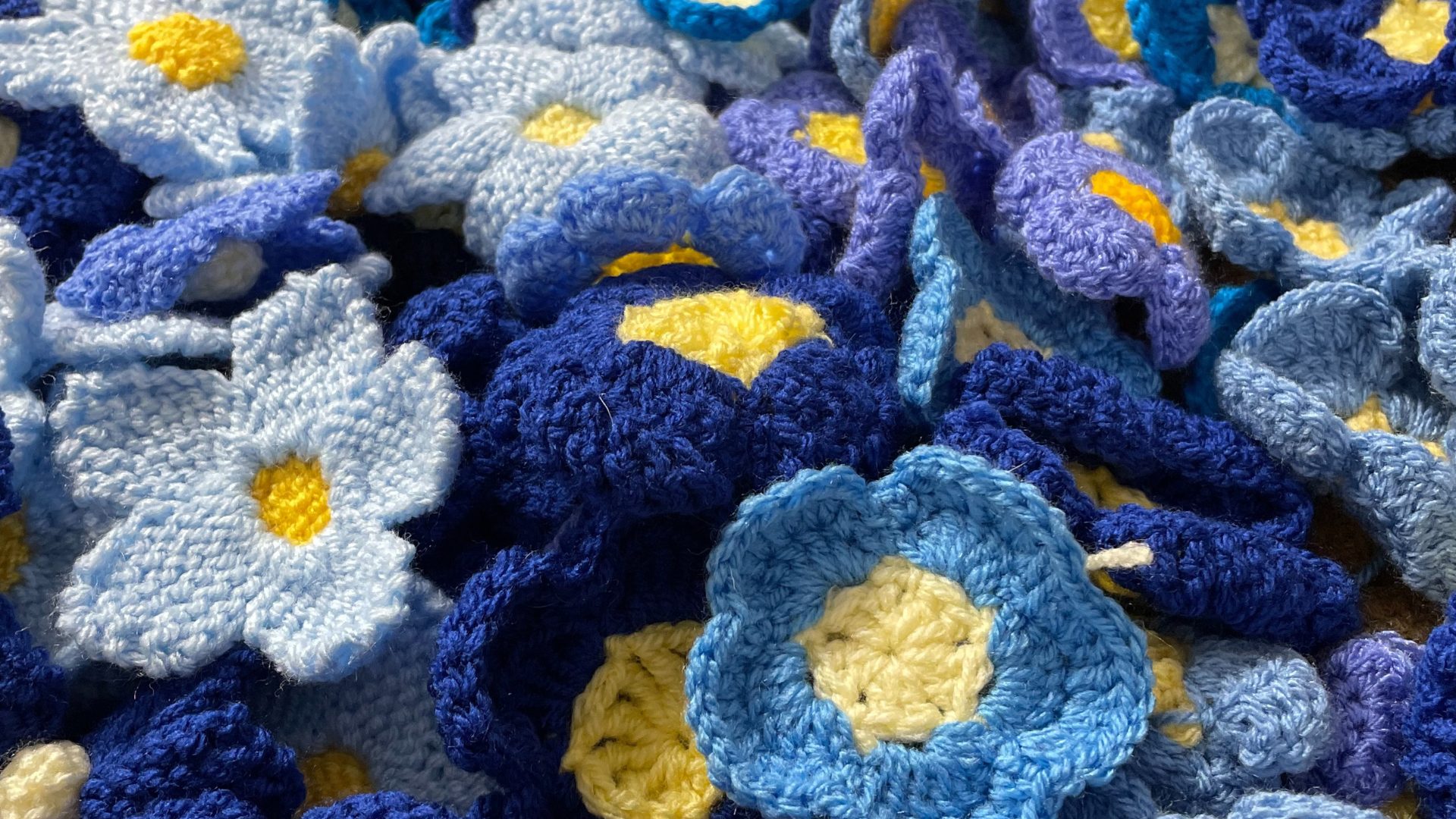 A collection of crocheted flowers in various shades of blue with yellow centres. The flowers are made of yarn and have a soft, textured appearance. The variety of blue hues—from light sky blue to deep navy—adds depth and a sense of richness to the composition. It's a lovely and cosy display of fibre art!