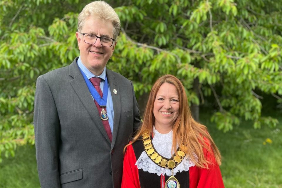 This photograph shows two individuals standing outdoors in front of a lush green background with trees. The man on the left is wearing a dark grey suit, a red tie, and glasses. He has short, light-colored hair and is smiling. He is also wearing a ceremonial medallion on a blue ribbon around his neck. The woman on the right is wearing formal ceremonial attire, which includes a red robe, a white lace jabot (a decorative frill), and a large gold ceremonial chain around her neck with a medallion. She has long, reddish-brown hair and is also smiling. Her outfit suggests she may hold a formal civic position, such as a mayor or council official. The setting and attire suggest this is a formal or official occasion, possibly related to a civic event or public office ceremony.