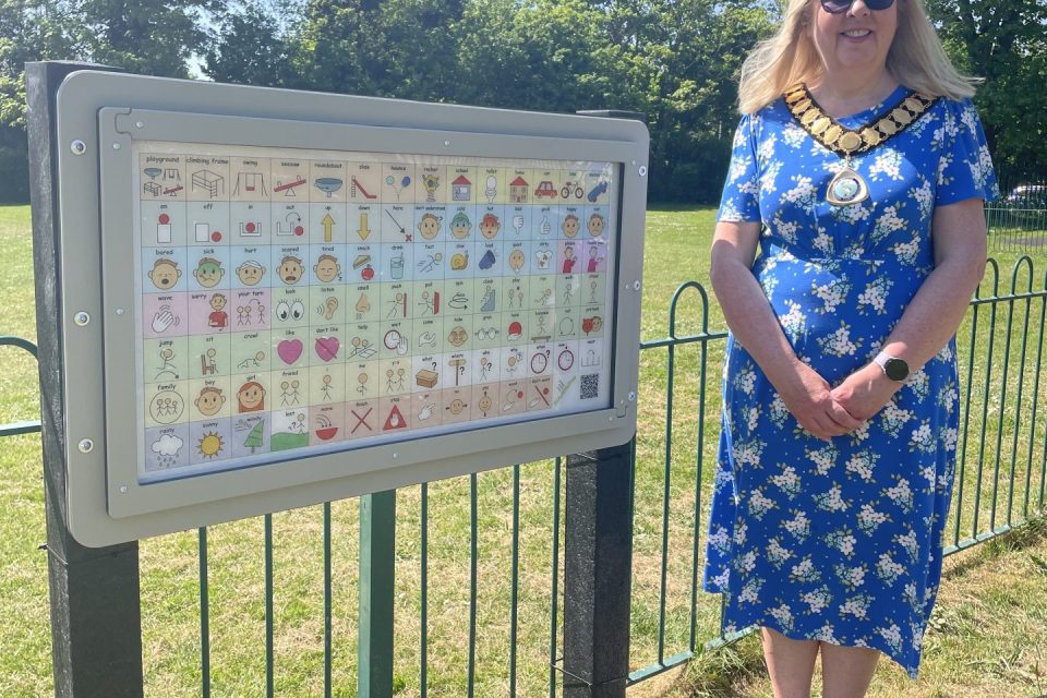 The image shows The Samoyor of Thatcham standing outdoors next to a large communication board mounted on a fence. She is wearing a bright blue floral dress, white sneakers, sunglasses, and a ceremonial gold chain. The background is a sunny park with green grass, trees, and a clear blue sky. The communication board is filled with symbols and icons, to help non-verbal individuals communicate. The board features various categories, such as emotions, activities, and objects typically found in a playground setting.
