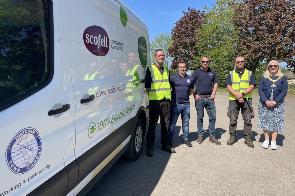 It shows a group of five people standing outdoors on a sunny day next to a white van. The van is branded with logos including "Scofell Commercial Landscapes" and "Thatcham Town Council," along with the text "100% Electric Vehicle." Two of the people are wearing high-visibility vests, two others are in navy blue polo shirts, and The Mayor is in a patterned dress and light jacket, wearing sunglasses and a ceremonial chain. In the background, there are trees with green and red foliage under a clear blue sky.