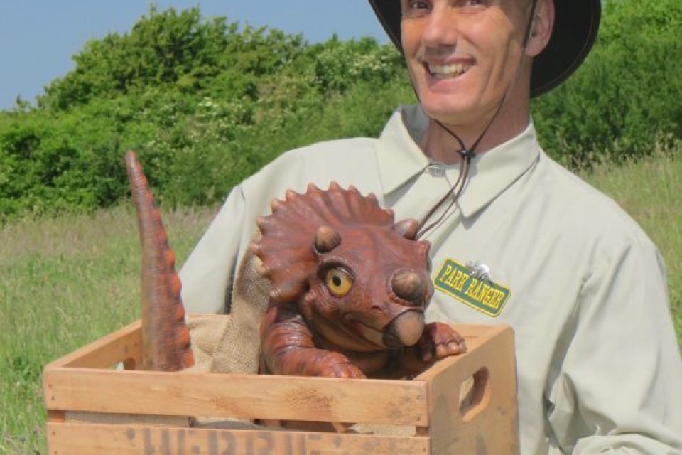A man holding a baby triceratops in a box. He is smiling broadly with a hat on, and a shirt with a Park Ranger tag
