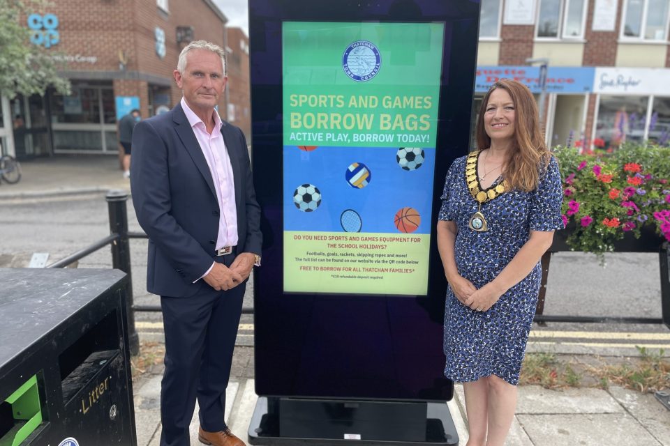 The image shows two individuals standing on either side of a new digital screen noticeboard in Thatcham Broadway. The person on the left is a man wearing a dark suit with a light pink shirt and brown shoes, standing with hands clasped in front of him. The person on the right is a woman wearing a blue patterned dress and gold mayoral chain, standing with hands clasped, smiling. The screen between them displays a colourful notice titled: “SPORTS AND GAMES BORROW BAGS – ACTIVE PLAY, BORROW TODAY!” It includes images of a football, volleyball, tennis racket, and basketball, with information about borrowing sports and games equipment for free for Thatcham families, along with a QR code for more information. This image captures the launch of the new digital screen noticeboard in the town centre, emphasising community initiatives and promoting active play for local families. Let me know if you need this formatted for Facebook with alt text or a caption for accessibility.