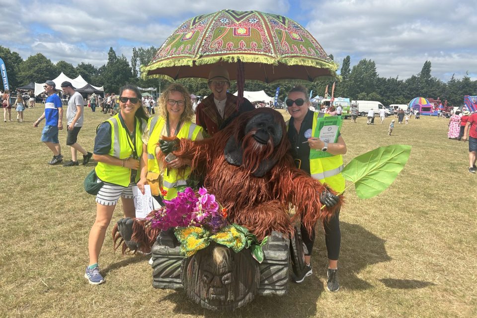Three members of the Thatcham Town Council team wear yellow high vis jackets and smiling broadly as an orangutang sits on a buggy between them underneath a parasol. It is a very sunny day,