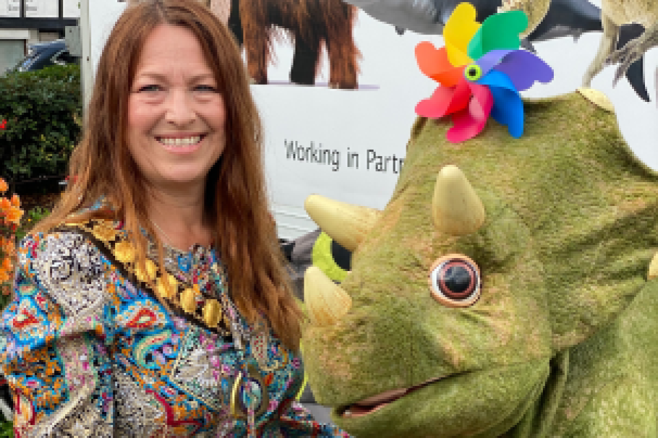 The image shows Mayor Councillor Pemberton standing next to a model of a triceratops. The dinosaur has three horns, large round eyes, and is wearing a colourful pinwheel decoration on its head. Cllr Pemberton is wearing a patterned, colourful top and Mayoral chains. Behind them is a banner with partial text and images of other prehistoric animals.