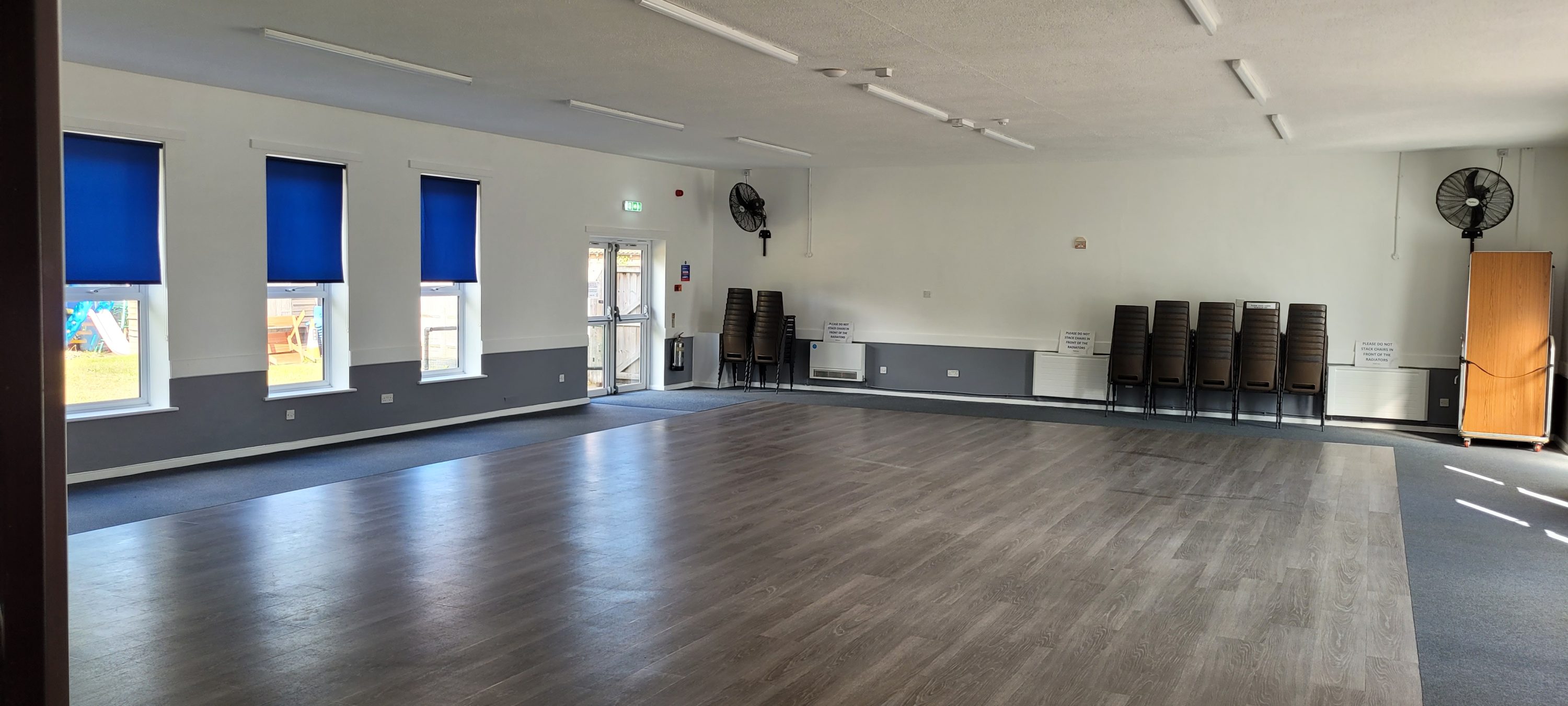 The image shows a clean, well-lit hall or community space. The walls are painted in two tones, white on top and gray on the bottom. The flooring appears to be a combination of wood-look planks in the center and carpet around the edges. Windows with blue roller blinds are visible along one wall, and there are stacks of chairs against the opposite wall. A couple of wall-mounted fans and signage regarding stacking chairs near radiators are also present. The space appears to be empty and ready for use, with a neutral and functional aesthetic.