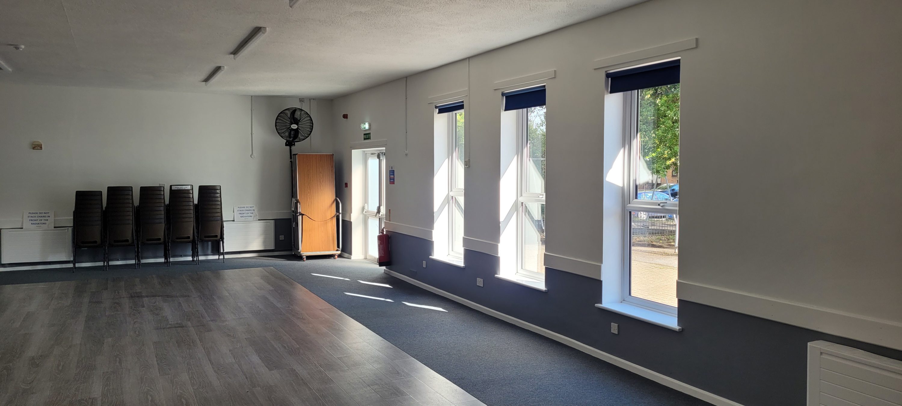 The image shows a brightly lit, empty hall. On the left, there are five stacked chairs next to a sign reminding people not to stack chairs in front of the radiators. A large fan stands near an exit door, accompanied by a table on wheels and a fire extinguisher. The right side features three tall windows through which sunlight streams, creating bright patches on the floor. The lower part of the walls is painted gray, while the upper portion is white. The floor is a combination of wood-patterned laminate and gray carpet. The space appears clean and well-maintained.